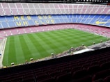 A large football stadium with empty seats arranged in vibrant blue and maroon colors. The well-maintained green field is visible, and a small group of people are gathered near one side of the field. The phrase 'MÉS QUE UN CLUB' is prominently displayed in yellow on the seats.