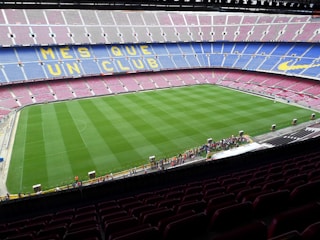 A large football stadium with empty seats arranged in vibrant blue and maroon colors. The well-maintained green field is visible, and a small group of people are gathered near one side of the field. The phrase 'MÉS QUE UN CLUB' is prominently displayed in yellow on the seats.