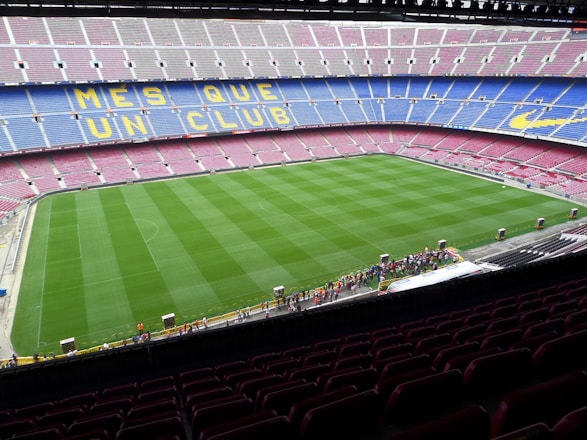A large football stadium with empty seats arranged in vibrant blue and maroon colors. The well-maintained green field is visible, and a small group of people are gathered near one side of the field. The phrase 'MÉS QUE UN CLUB' is prominently displayed in yellow on the seats.