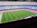 A large football stadium with empty seats arranged in vibrant blue and maroon colors. The well-maintained green field is visible, and a small group of people are gathered near one side of the field. The phrase 'MÉS QUE UN CLUB' is prominently displayed in yellow on the seats.