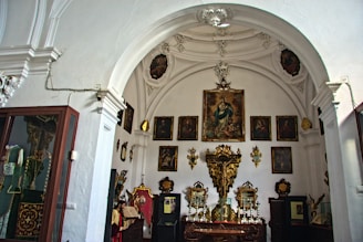 An ornate room with white arches and several religious paintings hanging on the walls. The focal point is a large painting in the center above an ornately decorated altar with gold accents. Various smaller objects and artifacts are displayed throughout the room, adding to the historical and sacred atmosphere.