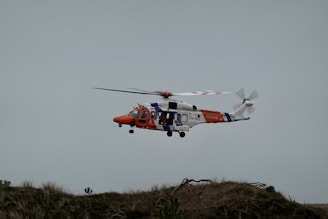 A rescue helicopter is flying above a grassy terrain. The helicopter is primarily white, orange, and blue, with 'RESCUE' and 'COAST GUARD' markings visible on its side.