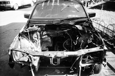 A black and white photo of a car missing its front parts, revealing the engine bay and various mechanical components. There are tools on the windshield and the scene is set in a driveway with a fence and another vehicle in the background.