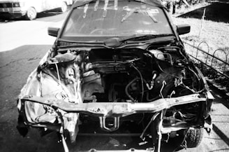 A black and white photo of a car missing its front parts, revealing the engine bay and various mechanical components. There are tools on the windshield and the scene is set in a driveway with a fence and another vehicle in the background.