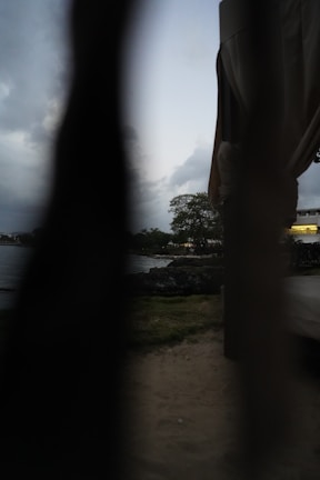A moody, dimly lit scene with partial view through a dark, narrow gap. The image features a seaside landscape with an expanse of water and cloudy sky, hinting at early evening or late afternoon. Faint outlines of trees and a structure are visible in the background.