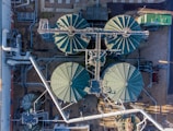 An aerial view of an industrial facility with large, circular tanks arranged in a structured layout. Metal pipes and walkways crisscross between the tanks, and there are smaller structures and equipment situated around them. The surrounding area includes gravel and some paved sections.