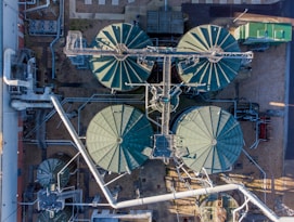 An aerial view of an industrial facility with large, circular tanks arranged in a structured layout. Metal pipes and walkways crisscross between the tanks, and there are smaller structures and equipment situated around them. The surrounding area includes gravel and some paved sections.