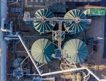 An aerial view of an industrial facility with large, circular tanks arranged in a structured layout. Metal pipes and walkways crisscross between the tanks, and there are smaller structures and equipment situated around them. The surrounding area includes gravel and some paved sections.