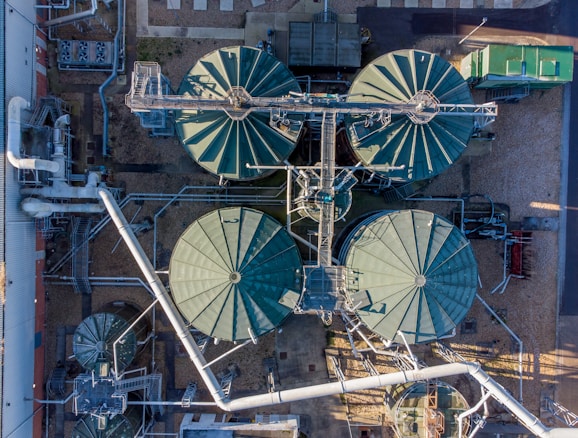 An aerial view of an industrial facility with large, circular tanks arranged in a structured layout. Metal pipes and walkways crisscross between the tanks, and there are smaller structures and equipment situated around them. The surrounding area includes gravel and some paved sections.