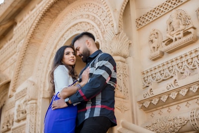 A couple stands closely together in front of an ornately carved stone wall. The wall features intricate patterns and sculptures, including figures in relief. The woman is wearing a blue dress and the man is in a plaid shirt. They are gazing into each other's eyes, suggesting a romantic moment.