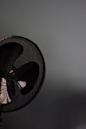 A close-up of a fan with a metal grill. The fan appears to be located against a plain, light-colored wall. The image focuses on the upper section of the fan, capturing the dark blades and the mesh pattern prominently.