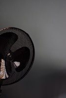A close-up of a fan with a metal grill. The fan appears to be located against a plain, light-colored wall. The image focuses on the upper section of the fan, capturing the dark blades and the mesh pattern prominently.