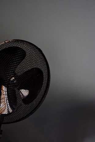 A close-up of a fan with a metal grill. The fan appears to be located against a plain, light-colored wall. The image focuses on the upper section of the fan, capturing the dark blades and the mesh pattern prominently.