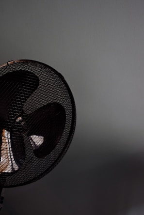 A close-up of a fan with a metal grill. The fan appears to be located against a plain, light-colored wall. The image focuses on the upper section of the fan, capturing the dark blades and the mesh pattern prominently.