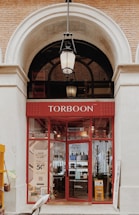 The image depicts the entrance of a store named Torboon with a red storefront and large glass windows displaying various items. A hanging lantern light is positioned above the entrance, framed by a large archway with exposed brick walls.
