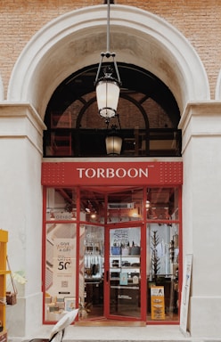 The image depicts the entrance of a store named Torboon with a red storefront and large glass windows displaying various items. A hanging lantern light is positioned above the entrance, framed by a large archway with exposed brick walls.