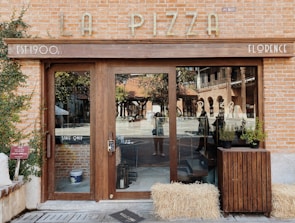 A rustic storefront with a wooden door and large windows displaying reflections of outdoor scenery. The name 'La Pizza' is displayed on a brick facade, and there are potted plants and hay bales near the entrance. The establishment is noted as being established in the 1900s and located in Florence.