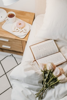 An open book rests on a neatly made bed with white sheets, accompanied by a bouquet of light pink roses. A wooden bedside table holds a cup of tea, a donut, and a decorative item spelling 'love.' The setting suggests a cozy and calm atmosphere.