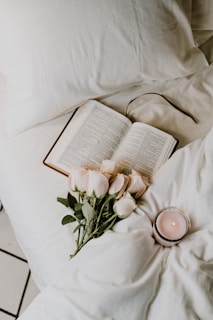 An open book lies on a cozy bed. Beside the book, there is a bouquet of light pink roses and a lit candle in a glass holder. The white bedding creates a soft and calm atmosphere.