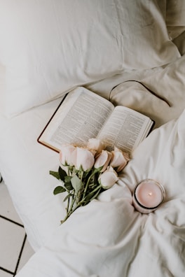 An open book lies on a cozy bed. Beside the book, there is a bouquet of light pink roses and a lit candle in a glass holder. The white bedding creates a soft and calm atmosphere.