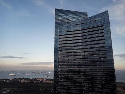 A modern high-rise building with a sleek, curved design stands prominently against a background of a clear sky. The building features many glass windows reflecting the surroundings. Below, there are several large ships dispersed across a wide body of water, suggesting a bustling port or harbor. The colors in the sky transition from a lighter blue to a soft orange, indicating either sunrise or sunset.