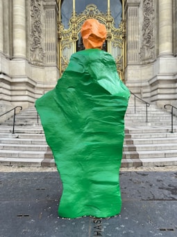 A large, abstract sculpture with a vibrant green body and an orange top stands in front of ornate, historic architecture. The sculpture is positioned on a set of stone steps leading to an intricate golden door, flanked by stone columns and detailed carvings on the walls.