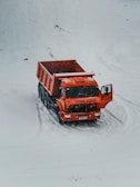 A bright orange dump truck is positioned on a snow-covered landscape with its door ajar. The vehicle stands out starkly against the white background, with light tire tracks visible in the snow around it.