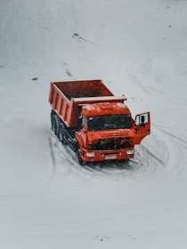 A bright orange dump truck is positioned on a snow-covered landscape with its door ajar. The vehicle stands out starkly against the white background, with light tire tracks visible in the snow around it.