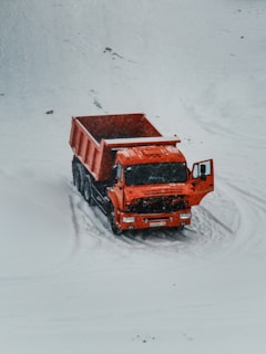A bright orange dump truck is positioned on a snow-covered landscape with its door ajar. The vehicle stands out starkly against the white background, with light tire tracks visible in the snow around it.