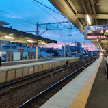A train station platform during twilight, with the sky displaying hues of blue and pink. The tracks run parallel to the platform, illuminated by overhead lights. A few people are present on the platform, and there are signs written in Japanese.