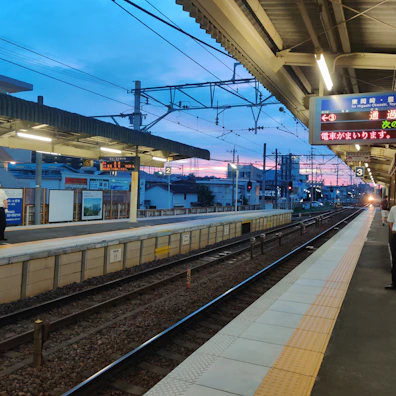 A train station platform during twilight, with the sky displaying hues of blue and pink. The tracks run parallel to the platform, illuminated by overhead lights. A few people are present on the platform, and there are signs written in Japanese.
