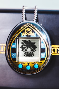 A handheld digital pet device features a pixelated character on its screen. The design includes a decorative gold and black frame with blue accents and buttons, and a chain attached at the top.