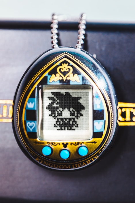 A handheld digital pet device features a pixelated character on its screen. The design includes a decorative gold and black frame with blue accents and buttons, and a chain attached at the top.