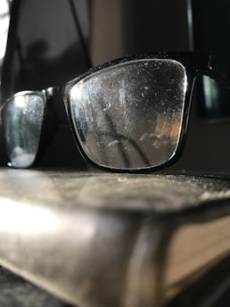 A close-up of a pair of glasses resting on an open book with a blurred background of a sunny park.