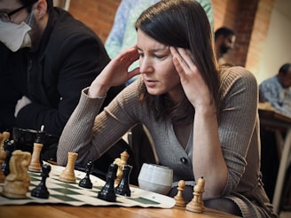 A person is seated at a chessboard, deeply concentrating on the game. Both hands are placed on the sides of their head, suggesting intense focus or contemplation. In the background, other people are present, with one person wearing a face mask. The setting appears casual, likely a café or a community gathering space.
