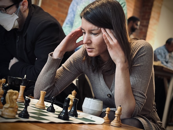 A person is seated at a chessboard, deeply concentrating on the game. Both hands are placed on the sides of their head, suggesting intense focus or contemplation. In the background, other people are present, with one person wearing a face mask. The setting appears casual, likely a café or a community gathering space.