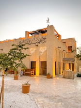 A traditional Middle Eastern building with Arabic calligraphy on the facade. The structure features earthy tones, with arched windows and wooden elements. A few wooden benches and potted plants are placed in the courtyard, which is paved with stone. A bird is seen flying above the building.