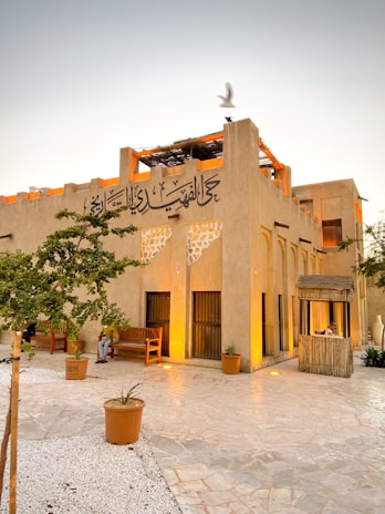 A traditional Middle Eastern building with Arabic calligraphy on the facade. The structure features earthy tones, with arched windows and wooden elements. A few wooden benches and potted plants are placed in the courtyard, which is paved with stone. A bird is seen flying above the building.