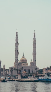 A large, ornate mosque with two tall minarets and a central dome is prominently featured near a waterfront. The structure is surrounded by a cityscape, with other buildings and trees visible. The water in the foreground reflects the architecture and boats can be seen near the shoreline.