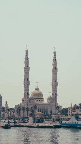 A large, ornate mosque with two tall minarets and a central dome is prominently featured near a waterfront. The structure is surrounded by a cityscape, with other buildings and trees visible. The water in the foreground reflects the architecture and boats can be seen near the shoreline.