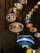 A series of intricately designed mosaic lamps hang from the ceiling, displaying vibrant patterns and colors. The lamps are suspended by dark chains, with each lamp featuring unique geometric designs comprised of multicolored glass tiles. Warm and cool hues intermingle, creating a dynamic visual display against a textured wall background.