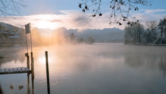 A calm lake at sunrise with mist rising over the water surrounded by pine trees.