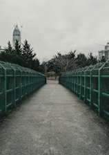 A concrete walkway flanked by green metal railings extends into the distance, bordered by trees on either side. A tall building and construction cranes are visible against a cloudy gray sky.