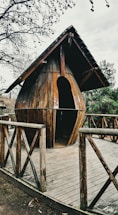 A wooden structure resembling a large water droplet serves as a small hut or shelter. It is placed on a wooden deck surrounded by a fence with cross beams. The setting is outdoors, and the trees nearby are bare and leafless, suggesting a winter or late autumn atmosphere.