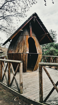 A wooden structure resembling a large water droplet serves as a small hut or shelter. It is placed on a wooden deck surrounded by a fence with cross beams. The setting is outdoors, and the trees nearby are bare and leafless, suggesting a winter or late autumn atmosphere.