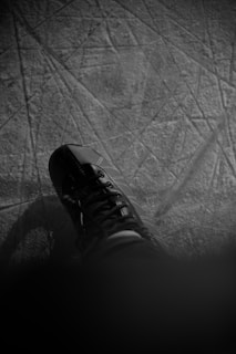 A black and white image of an ice skate on a textured ice surface. The skate is laced tightly and the toe section is prominently visible. The ice is marked with scratches and lines, suggesting recent activity.
