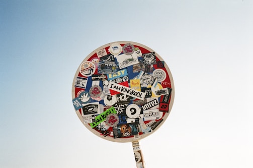 A circular sign is covered with a variety of colorful stickers and decals, each bearing different designs and text, standing out against a clear sky backdrop.