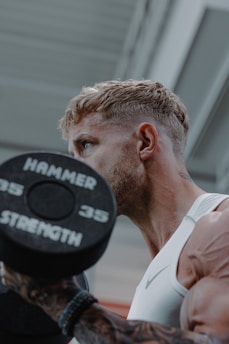 A person with short blond hair and tattoos is lifting a 35-pound dumbbell in a gym setting. The focus is on the person's muscular arm and the weight.