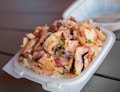 A dish featuring seasoned octopus pieces served over a bed of rice in a white disposable container. Garnished with green onions and spices, it appears fresh and appetizing.