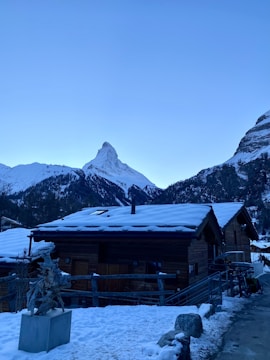 A picturesque winter scene featuring wooden chalets with snow-covered roofs in the foreground. In the background, a distinctive mountain peak rises against a clear blue sky. Snow blankets the surrounding landscape, and a pathway runs alongside the buildings.